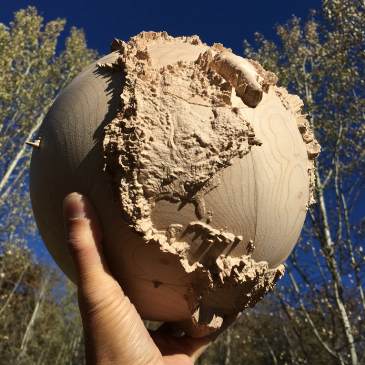 fully three-dimensional wood-carved terrestrial globe held in my hand with blue sky and trees in the background