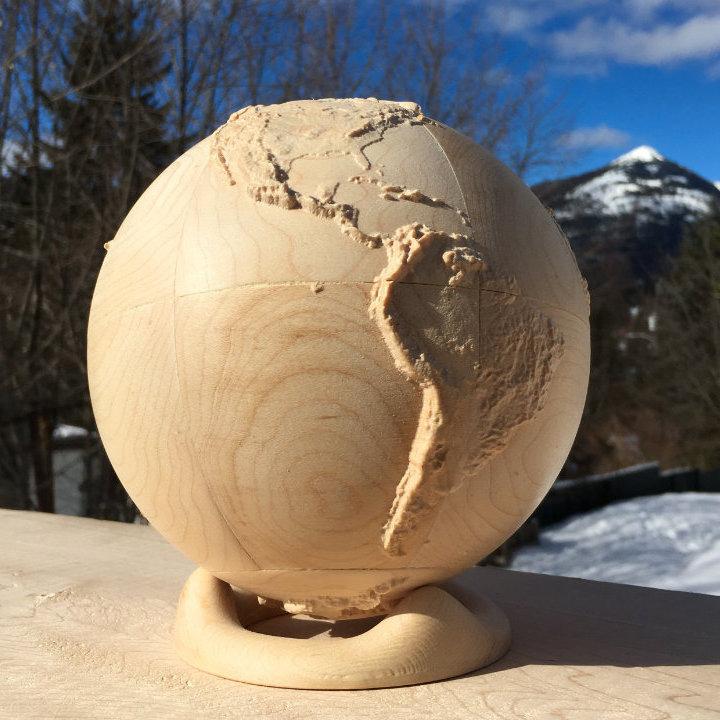fully three-dimensional wood-carved terrestrial globe on a simple stand with blue sky and mountains in the background
